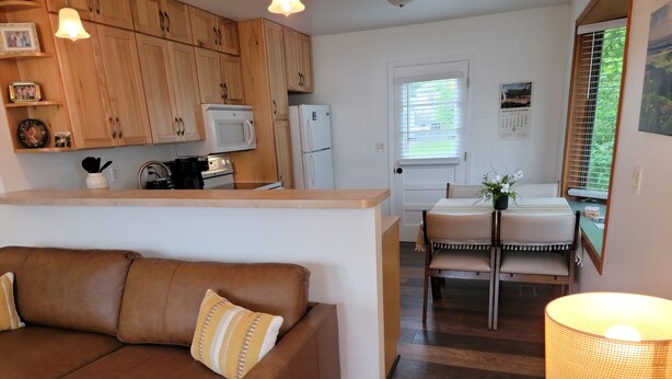 A cozy living room with a brown leather sofa and a wooden dining table with chairs. A kitchen with wooden cabinets and modern appliances is visible in the background.