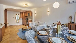 A modern dining area with a round table set for six, surrounded by upholstered chairs. A kitchen with wooden cabinets and a built-in oven is visible in the background. - Apartment in the Center of Paris (Paris)