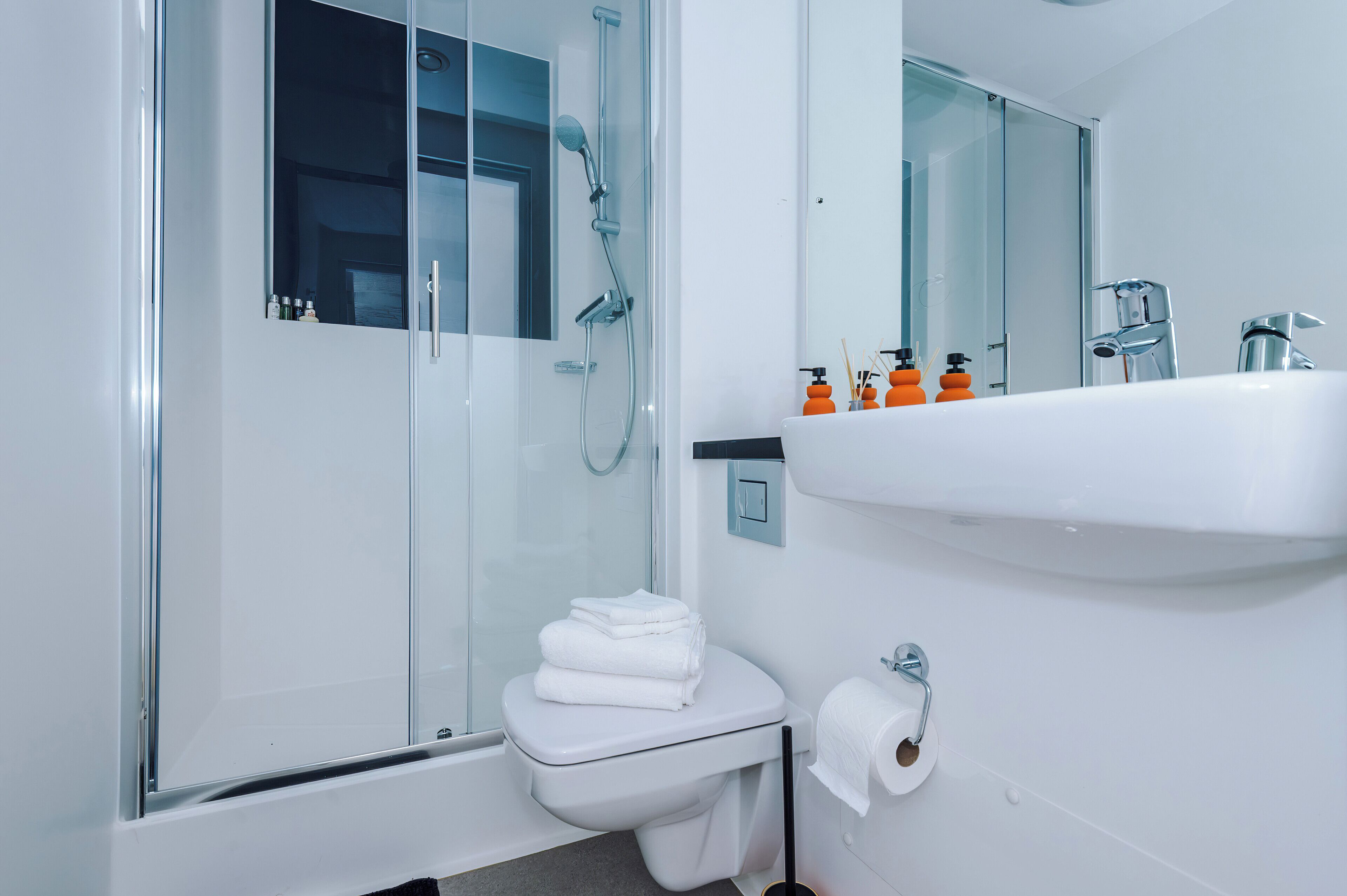 Un bany modern amb plat de dutxa envoltat de vidre, un vàter blanc i un lavabo amb productes de tocador.