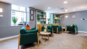 Un área de estar moderna con sofás verdes, una mesa de centro de madera y una estantería llena de libros y plantas.