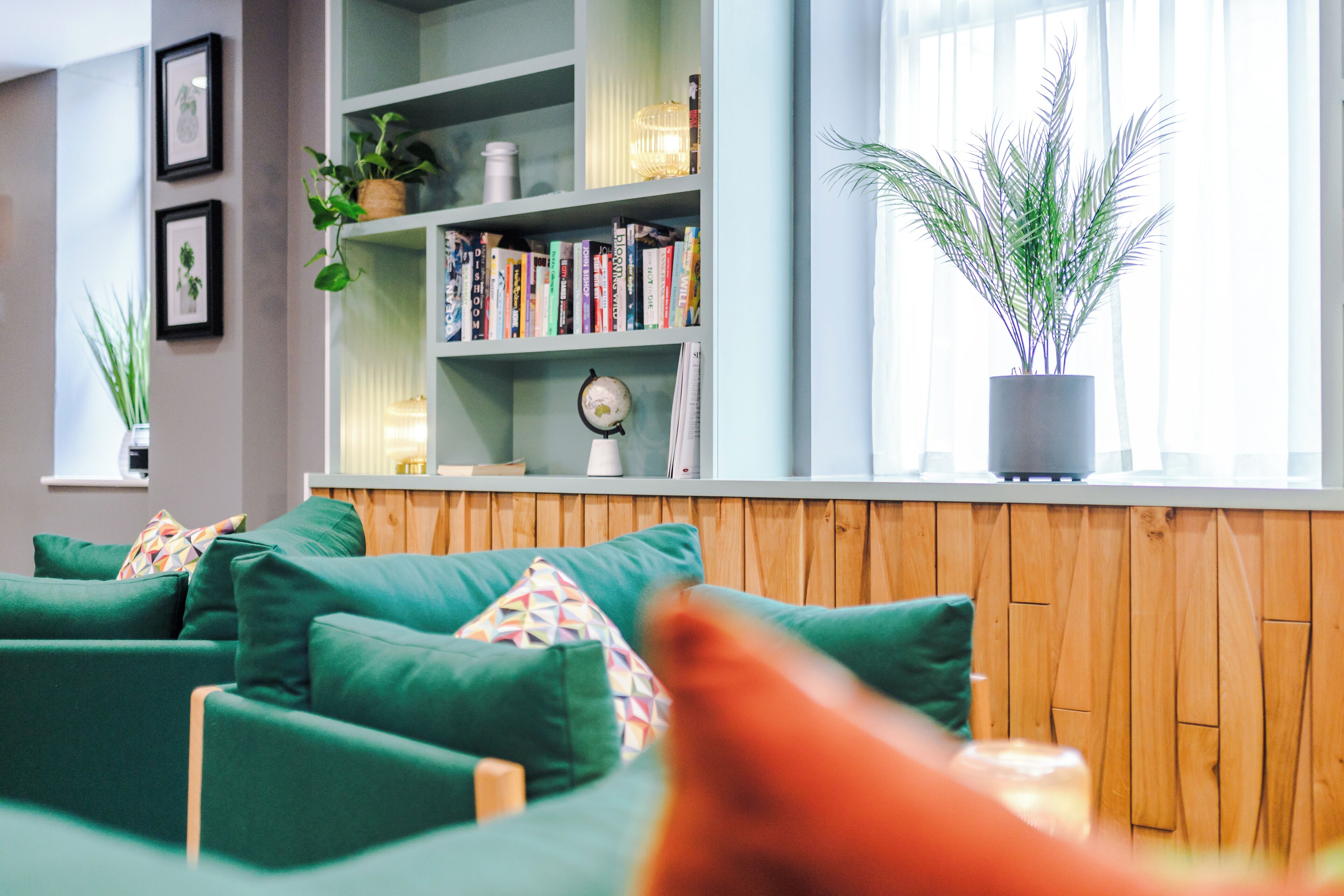 Un salon moderne avec un canapé vert, un panneau mural en bois, une bibliothèque remplie de livres et d’objets décoratifs, et une grande fenêtre ornée de rideaux légers.