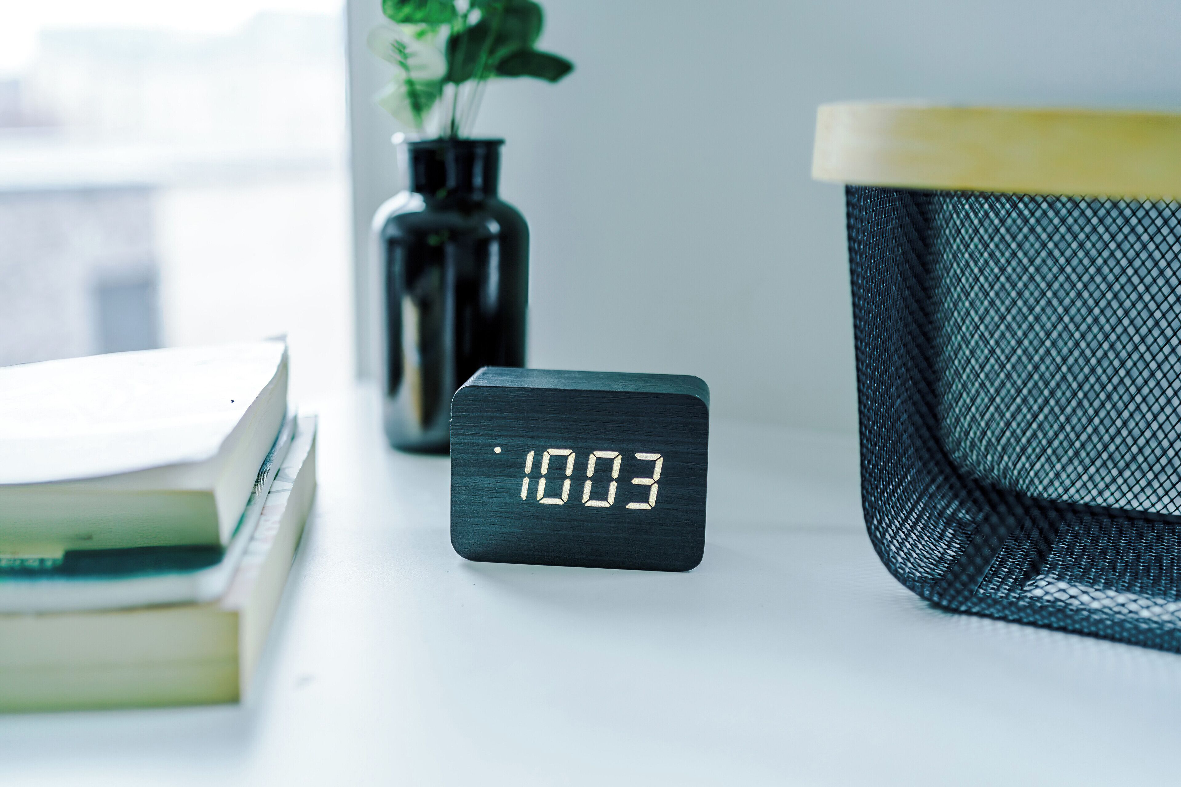 Une horloge numérique affichant 10h03, un vase avec une plante et une pile de livres sur une table.