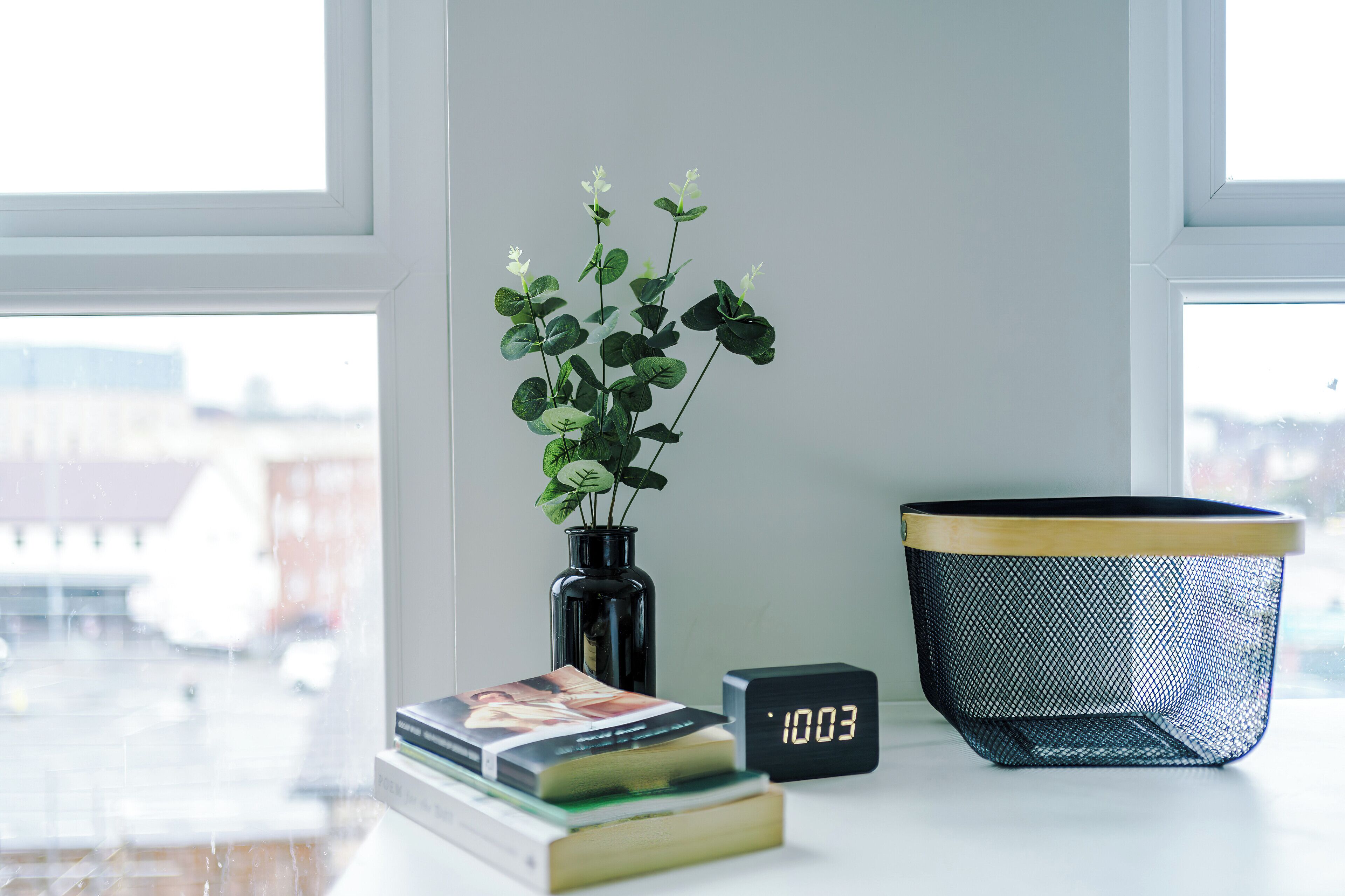 A window with a view of buildings, a black vase with white flowers, a stack of books, and a black digital clock displaying 1003.