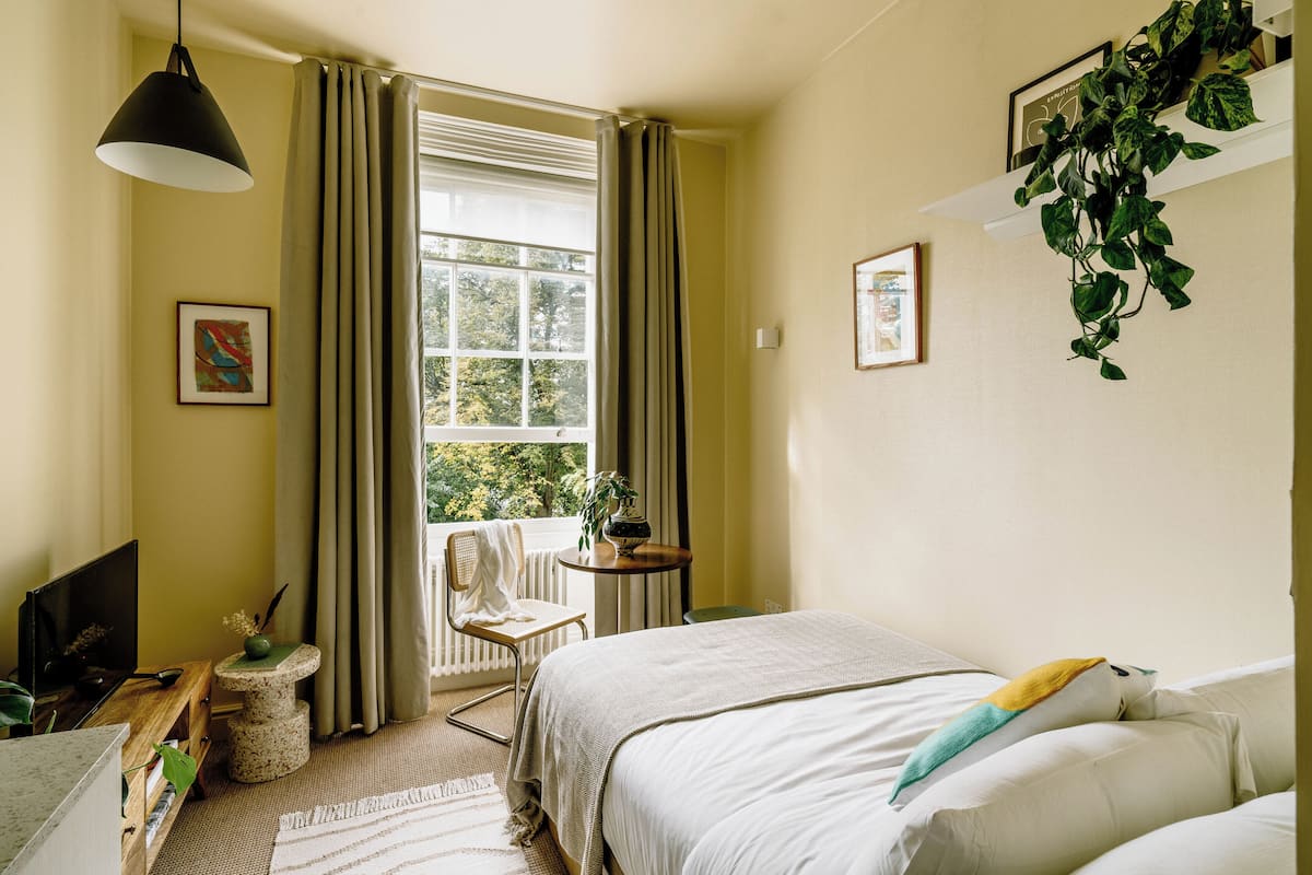 A bedroom with a bed, a chair, a small table, a TV, and a window with curtains.