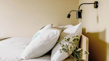 A neatly made bed with white bedding and pillows, a wall-mounted lamp, and a small vase with flowers.
