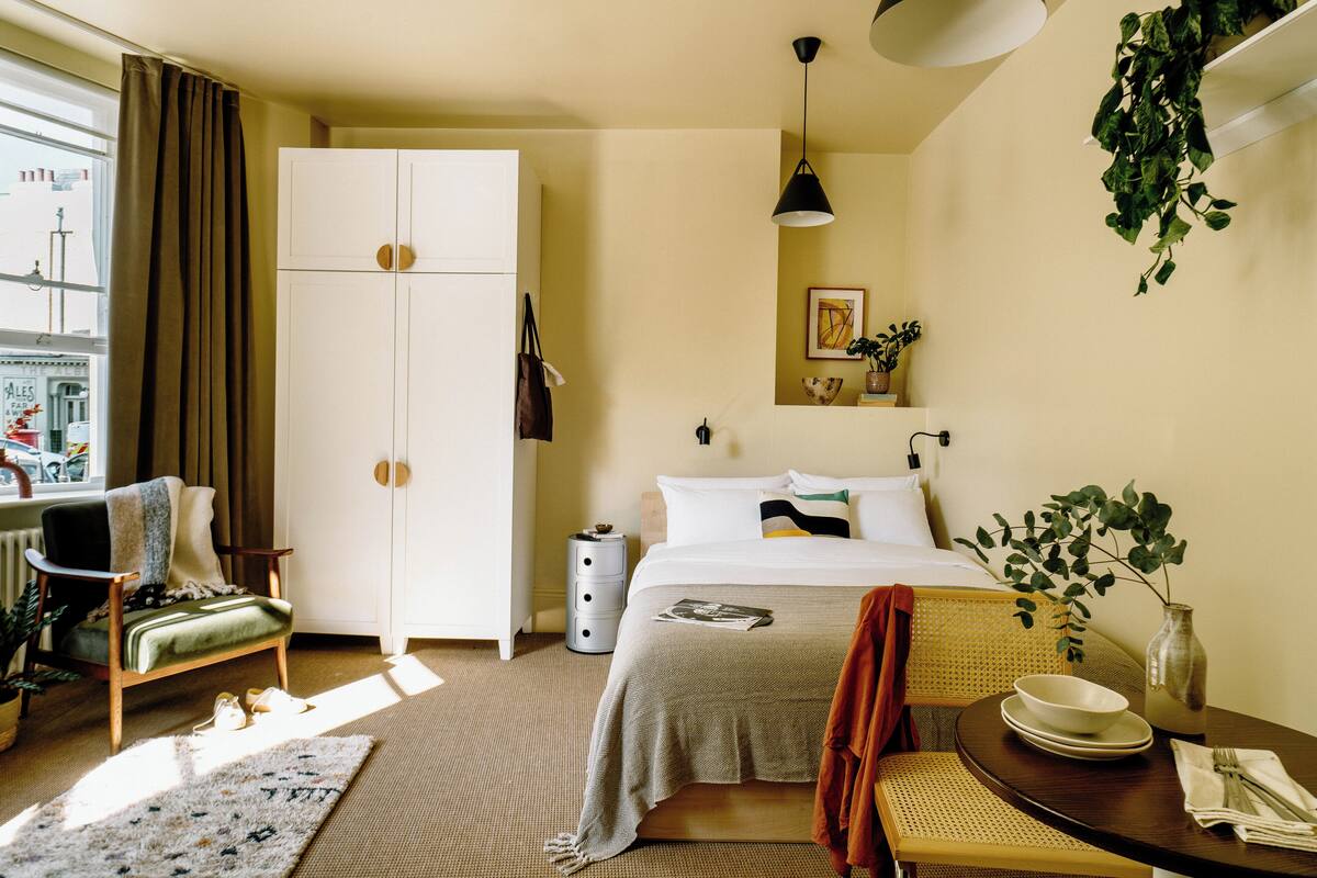 A hotel room with a bed, a chair, a desk, a wardrobe, a bedside table with a bowl and a vase, and a window with curtains.