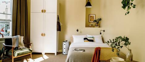 A hotel room with a bed, a chair, a desk, a wardrobe, a bedside table with a bowl and a vase, and a window with curtains.