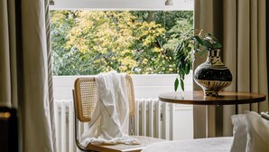 A bedroom with a bed, a chair, a small table with a vase, and a window with curtains. - Mason & Fifth, Primrose Hill (London)