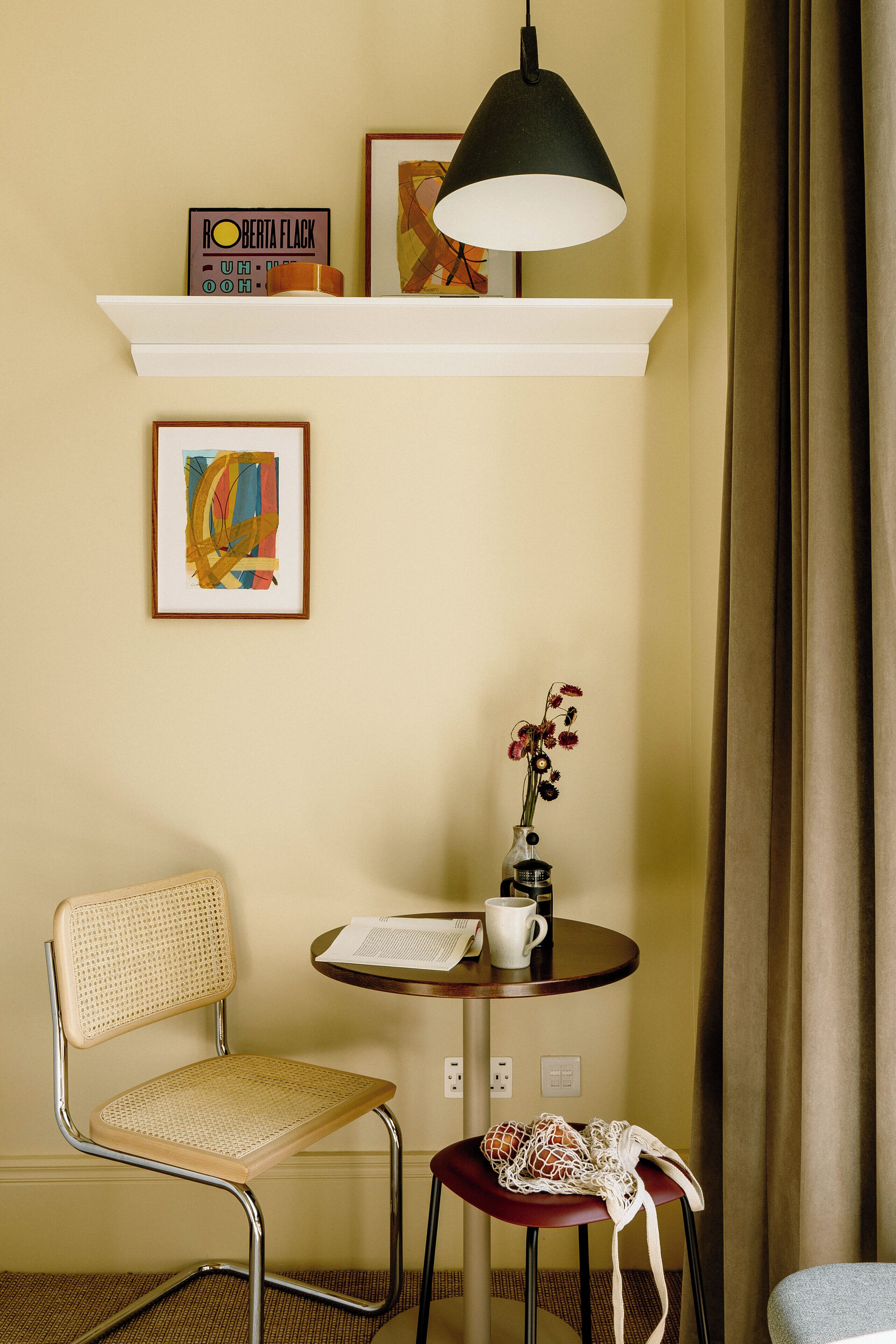 A small room with a white chair, a round table with a vase of flowers, a framed abstract painting, a shelf with a radio, and a hanging lamp.