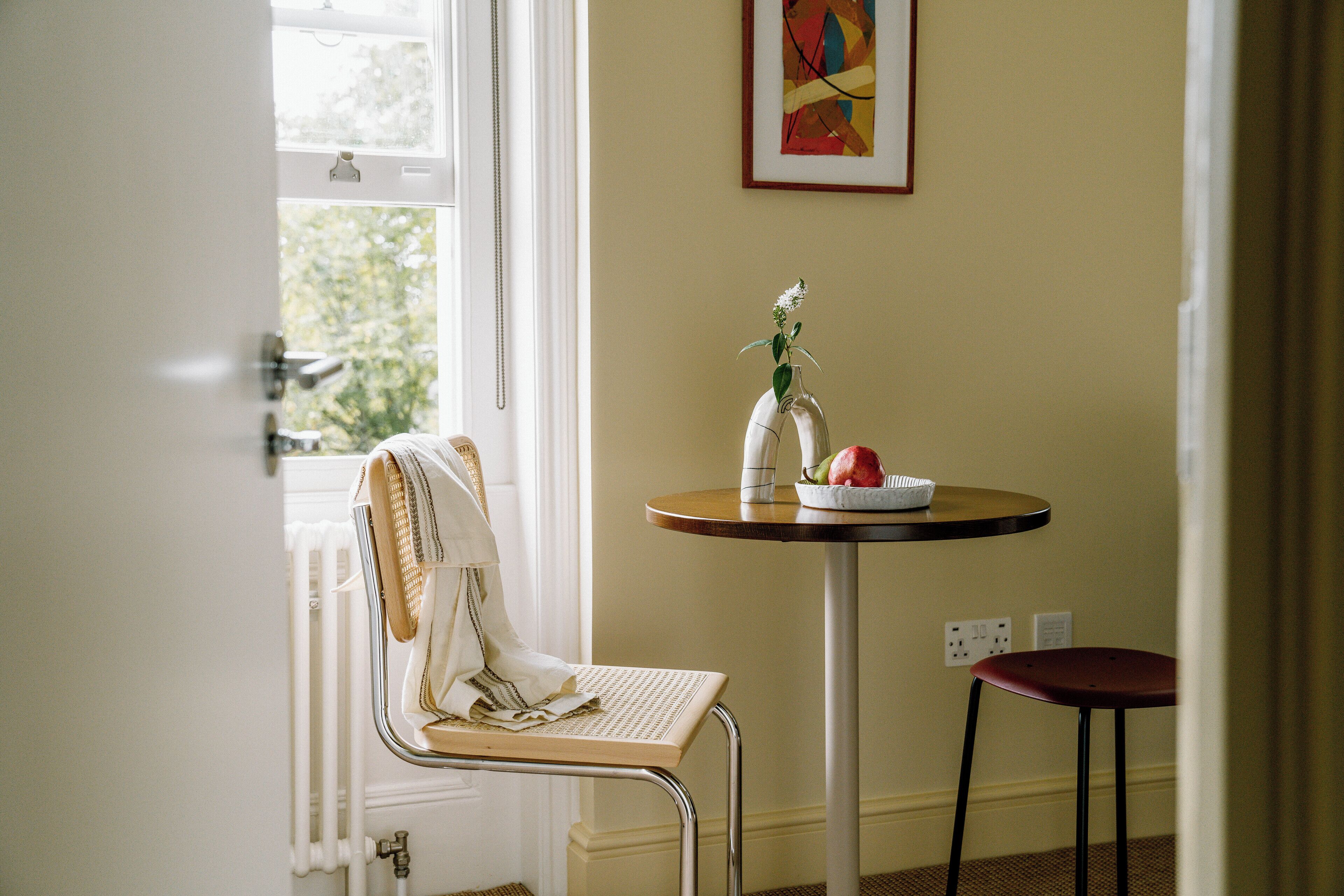 Une pièce avec un radiateur, une chaise, une table ornée d’un vase et de fruits, et un tableau accroché au mur.