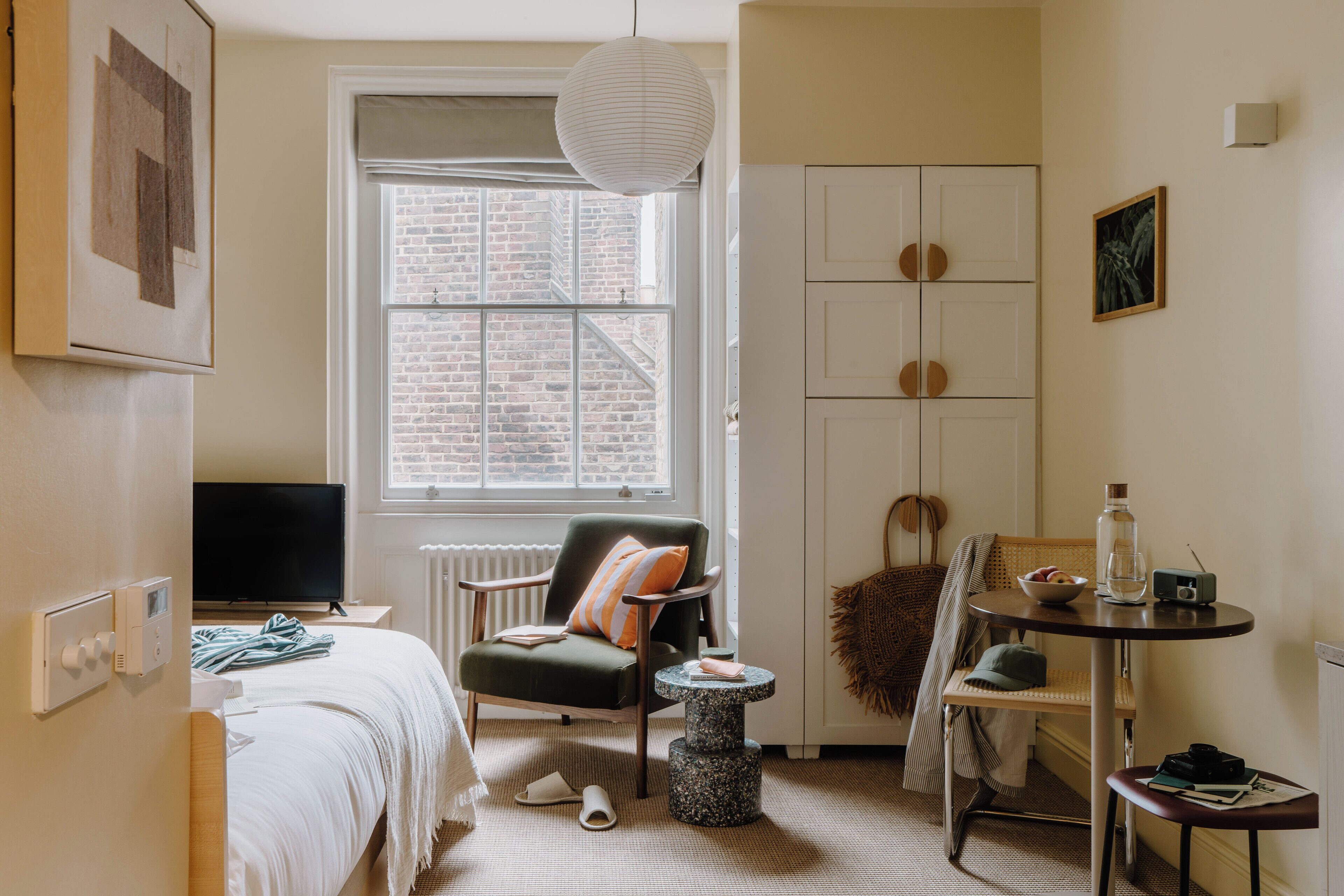 Habitación pequeña con cama, silla, mesita, radiador, ventana con vista a un edificio de ladrillo y una lámpara colgante.