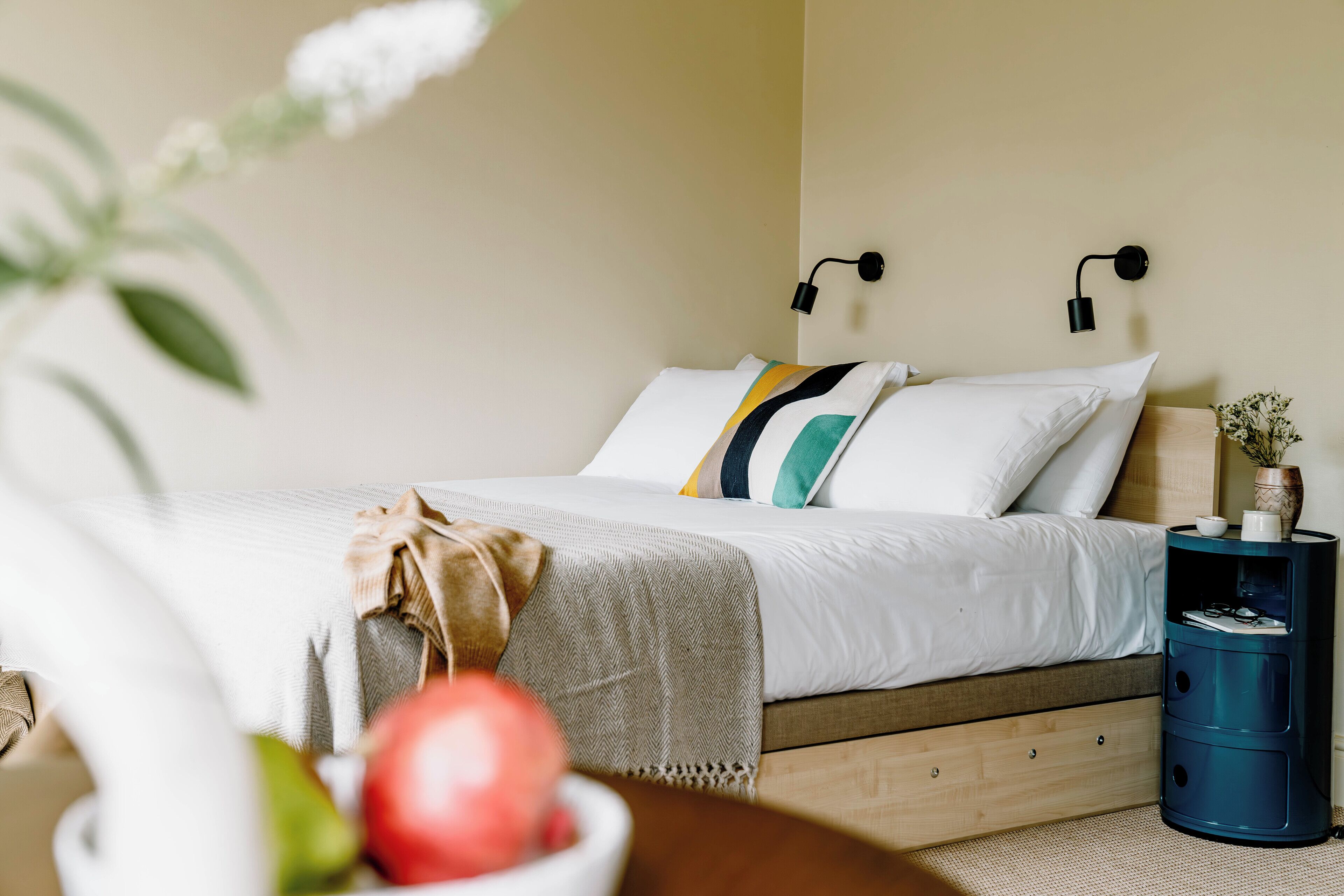 Un letto rifatto con cuscini bianchi e multicolore, una testata in legno e un comodino blu con un vaso e una ciotola di frutta.