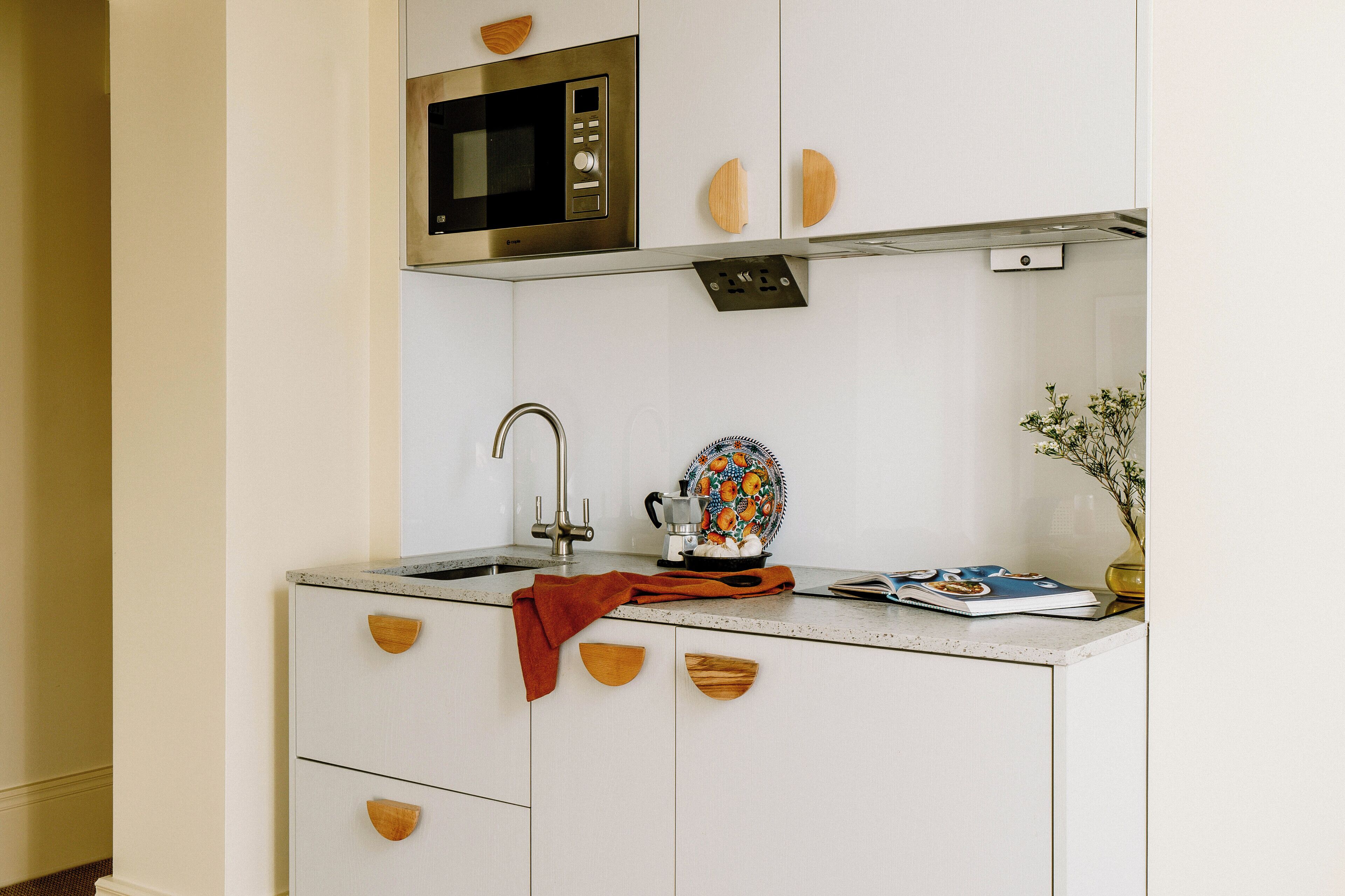 Uma cozinha compacta com micro-ondas integrado, pia e um balcão pequeno com um prato decorativo e uma revista.