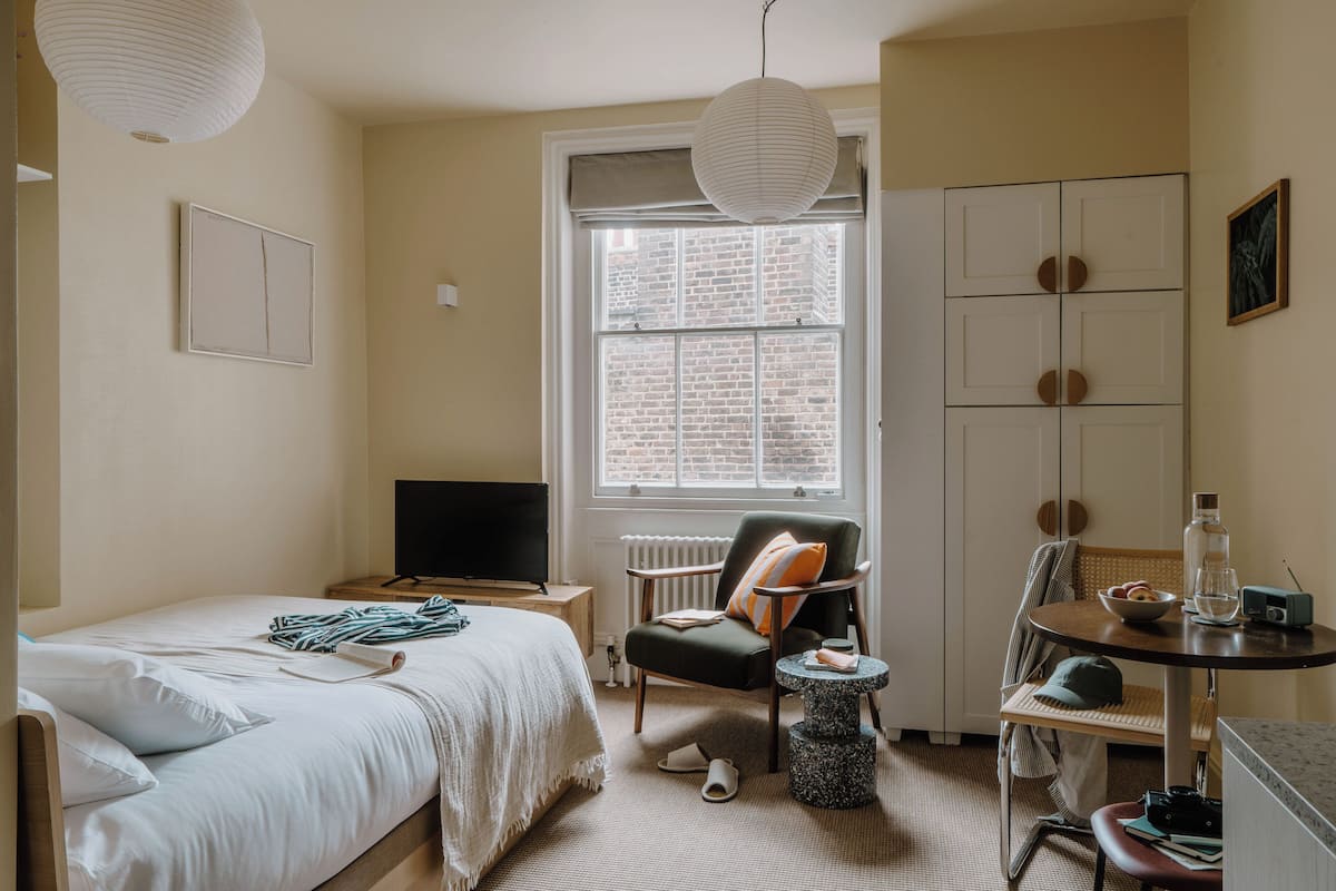 A bedroom with a bed, a chair, a table, a television, and a window with a view of a brick building.
