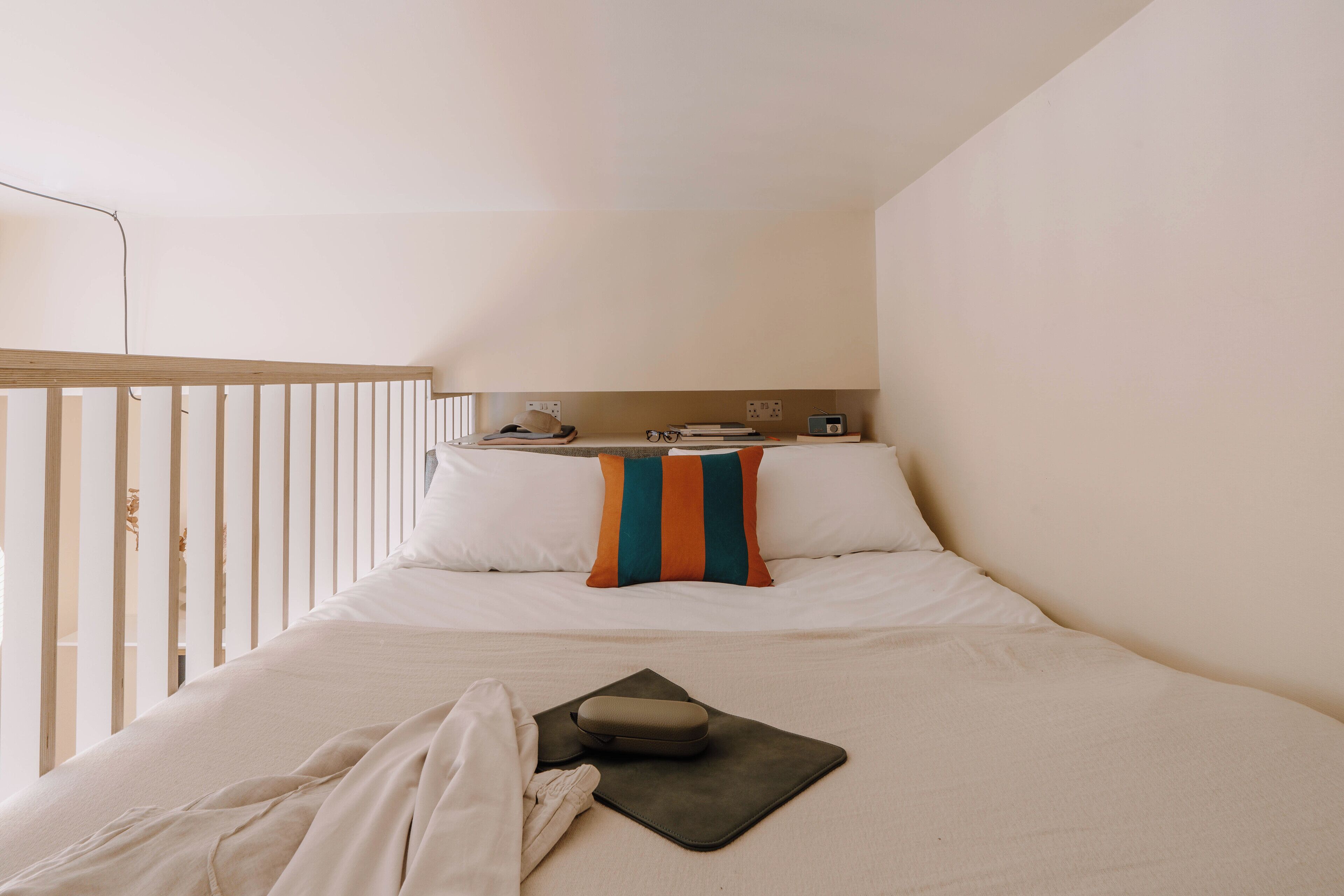 A hotel room with a bed, pillows, a bedside table with books and a radio, and a window with a view of the outside.