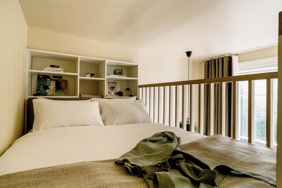 A neatly made bed with a patterned bedspread, a bedside shelf with books and a lamp, and a window with vertical blinds.