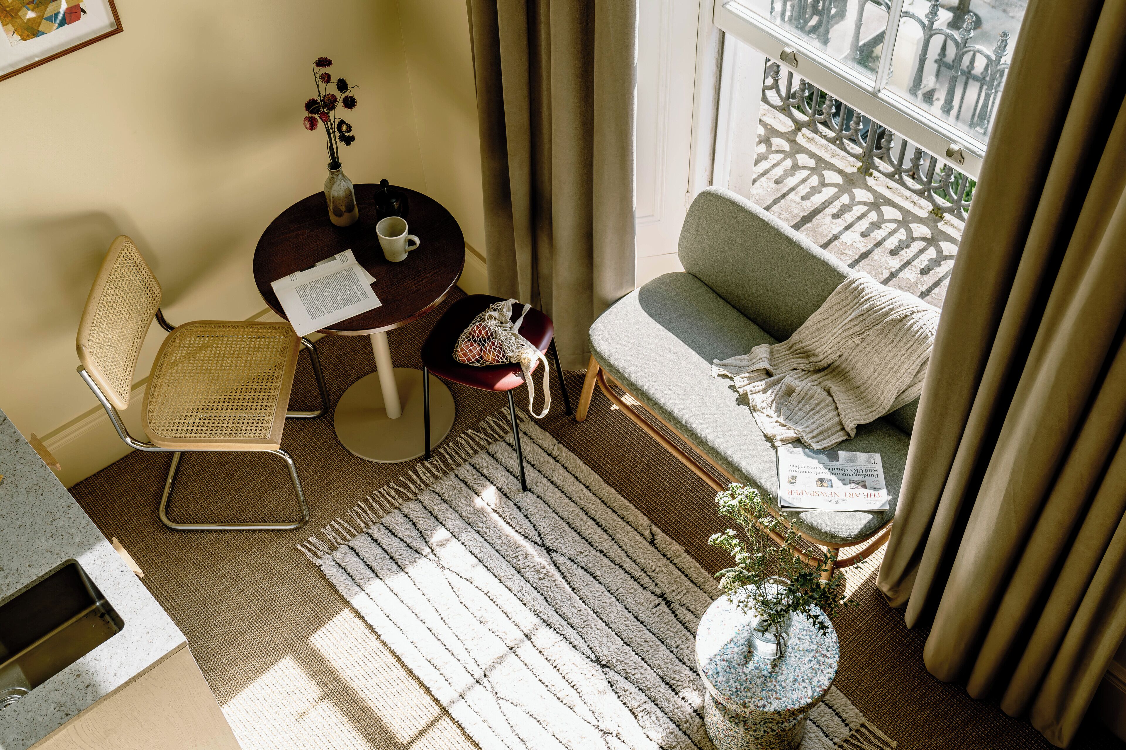 Une pièce confortable avec un canapé, un fauteuil, une petite table sur laquelle se trouvent un vase et des papiers, une fenêtre avec des rideaux et un balcon donnant sur un beau paysage.