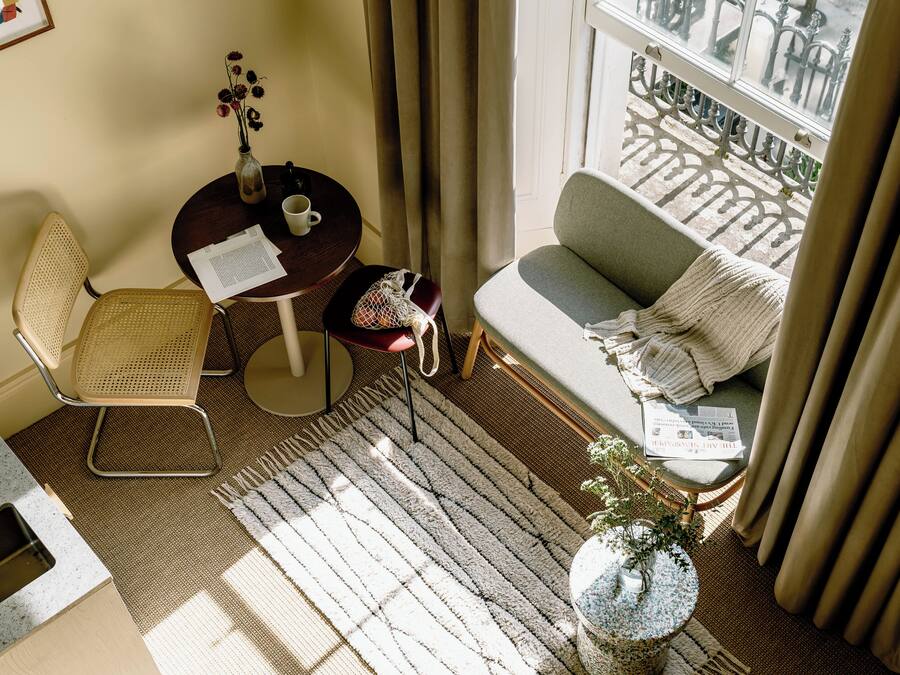 A cozy room with a sofa, a chair, a small table with a vase and papers, a window with curtains, and a balcony view.