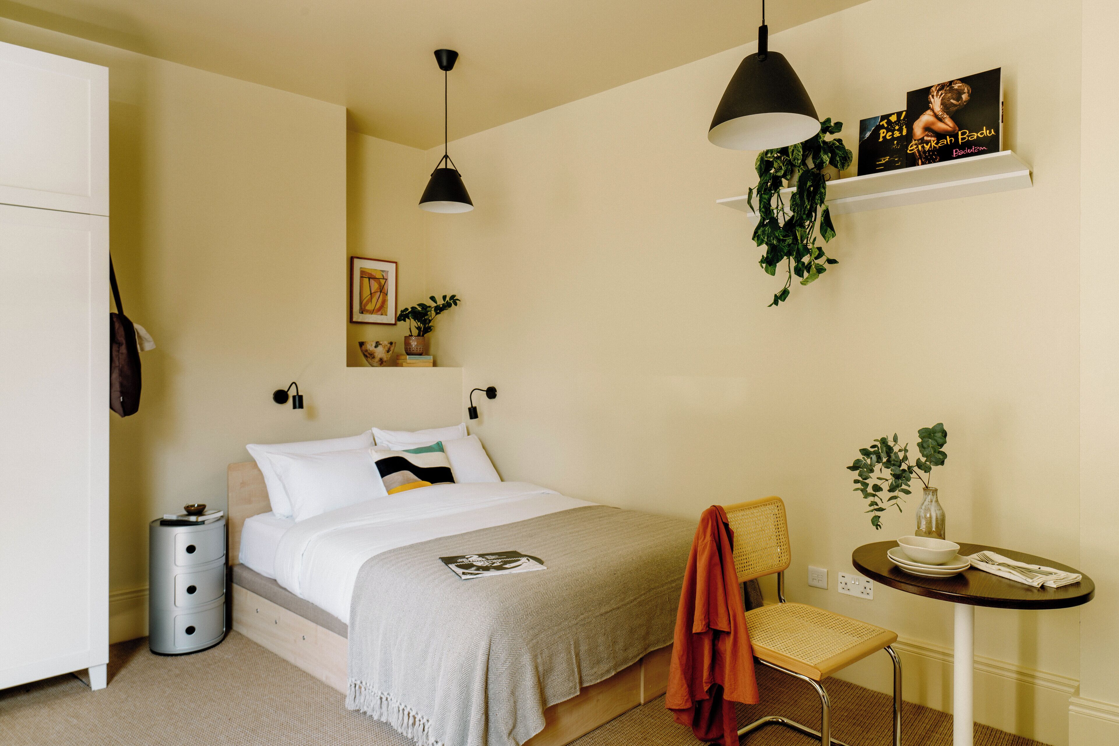 A neatly arranged bedroom with a bed, bedside table, chair, and a small table with a plant.