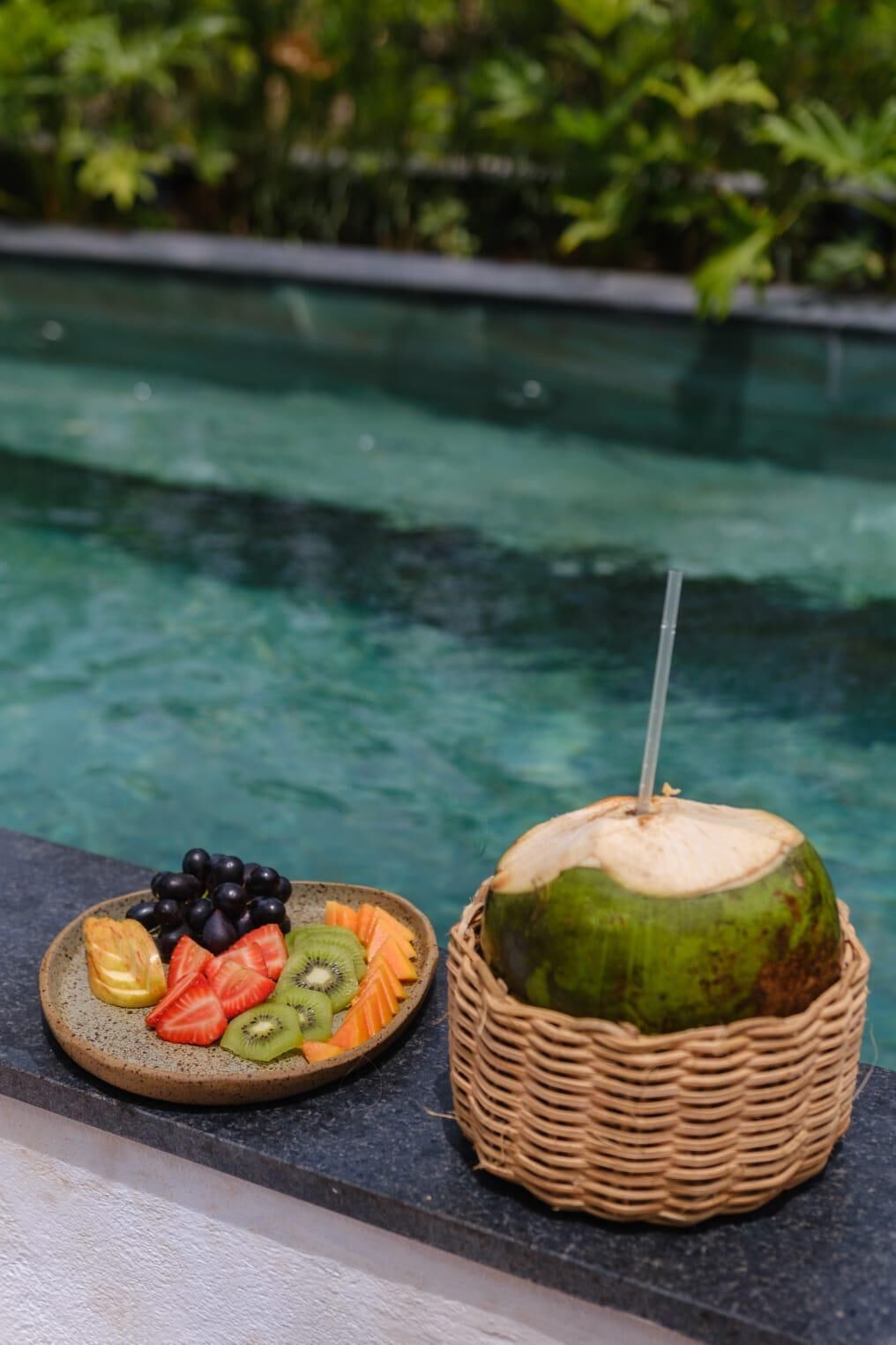 A plate of fruit and a coconut with a straw in a basket by a pool.