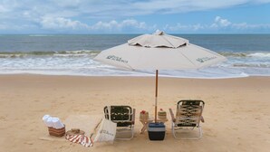 A beach setting with a beach umbrella, two beach chairs, a table with a basket and a small cake, and towels on the sand. - Vila Almesca (Porto Seguro)