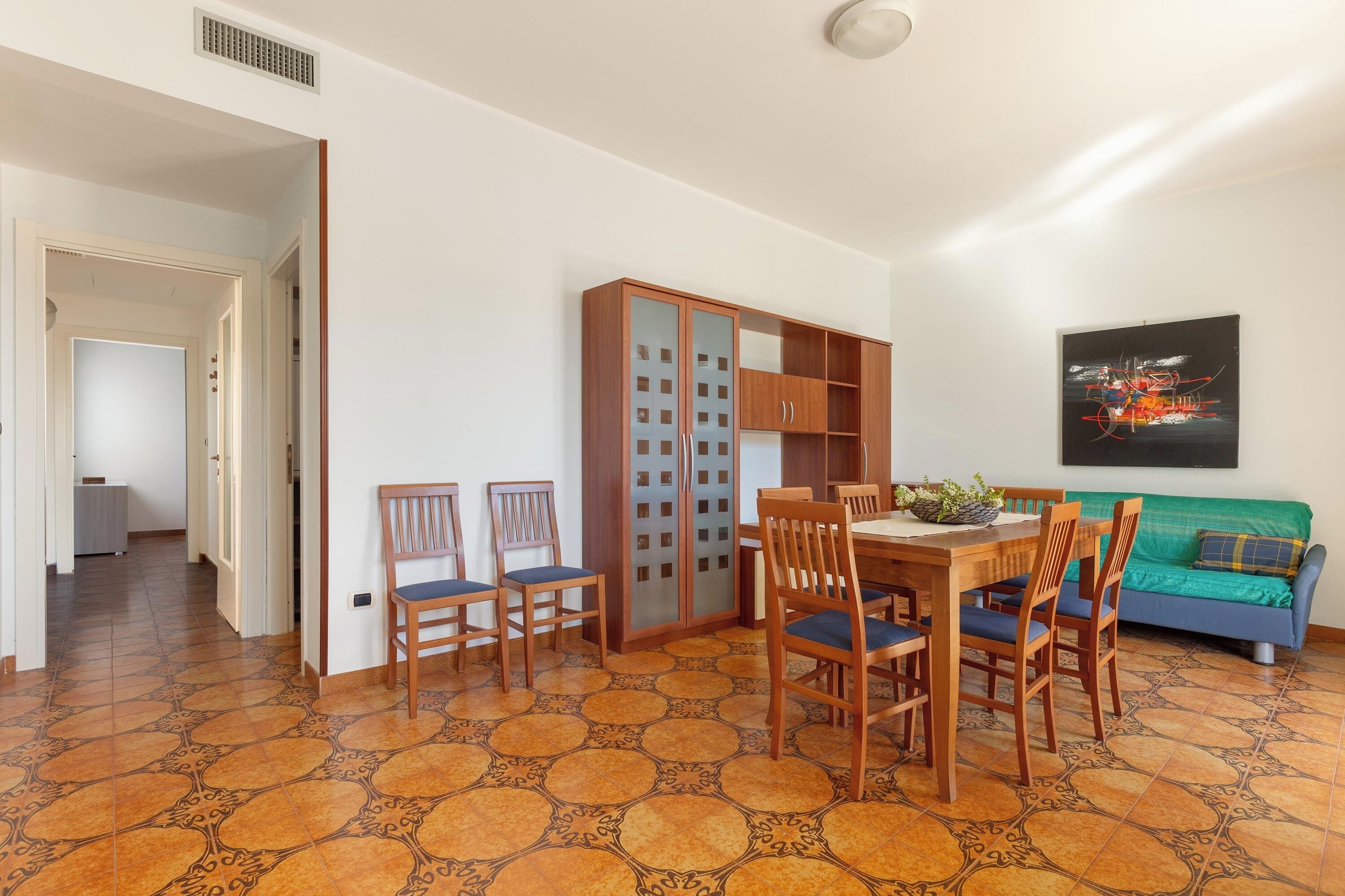 Un comedor con una mesa y sillas de madera, un mueble con puertas de vidrio y un sofá verde frente a una pared blanca.