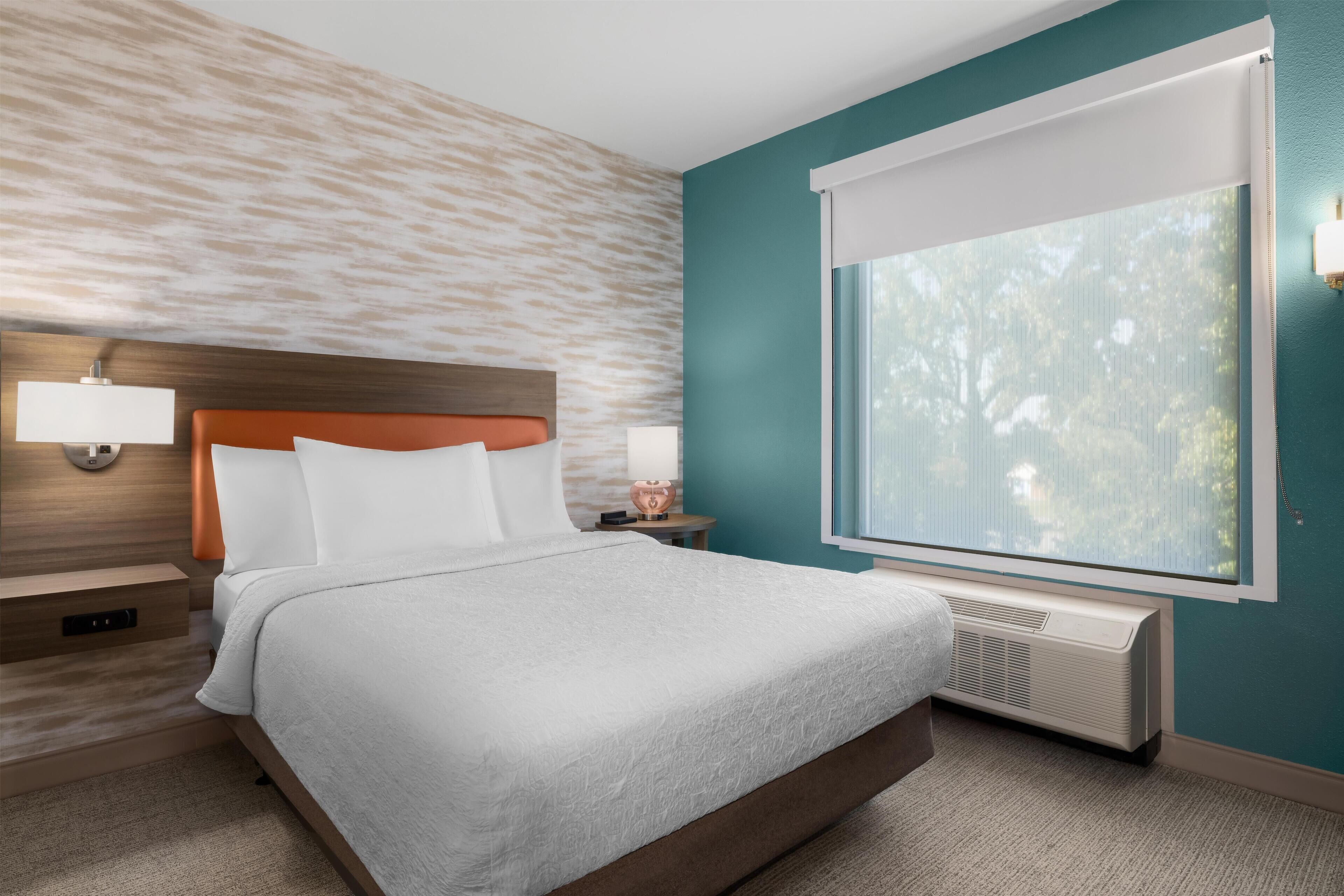 Habitación de hotel con cama, mesitas de noche, lámpara, ventana con persianas y un calefactor de pared.
