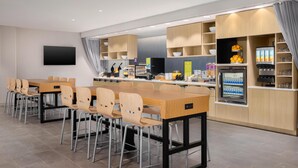 Un espace repas moderne avec une longue table en bois et des chaises, un téléviseur à écran plat fixé au mur et un réfrigérateur rempli de boissons.