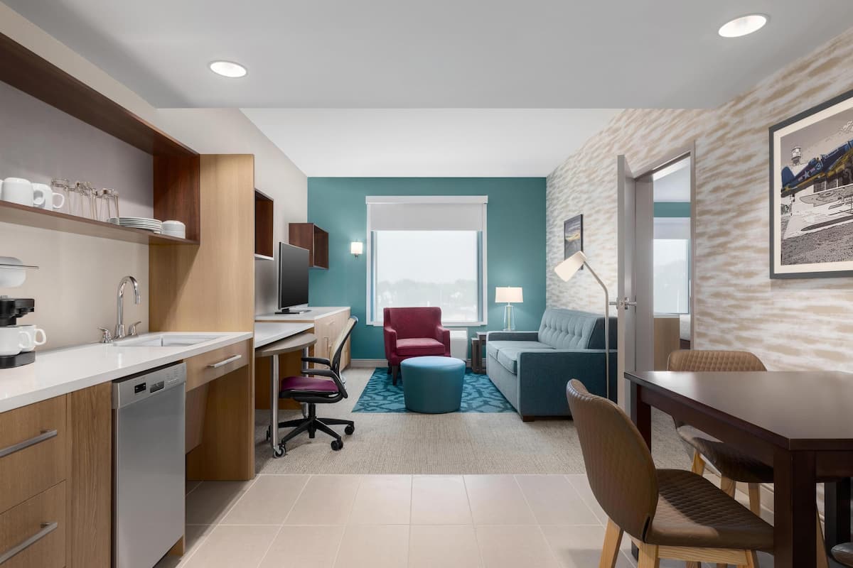 A modern kitchen with a dishwasher, a dining area with a table and chairs, a red chair, and a blue ottoman.