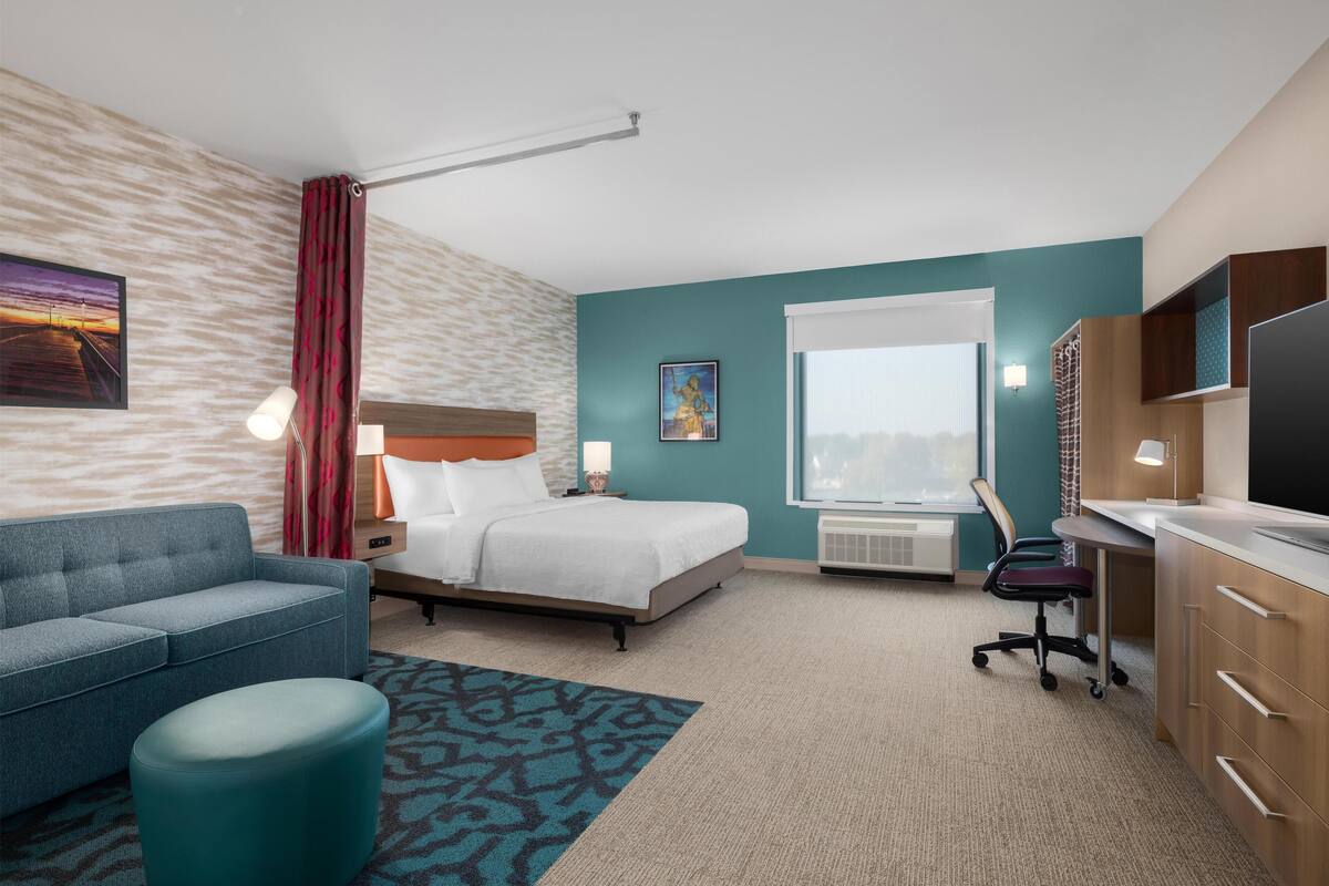A hotel room with a bed, a desk, a chair, a sofa, a TV, and a window.