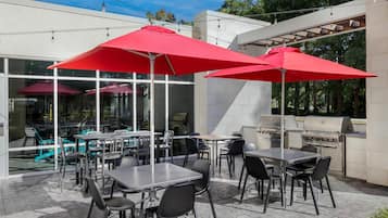 An outdoor patio area with tables and chairs, red umbrellas, and a barbecue grill.