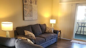 A living room with a grey sofa, two lamps, a ceiling fan, and a wall with a 'BEACH' sign. - 2103 Palms, 2-bedroom condo, Great Fall Weekends are still available (Rehoboth Beach)
