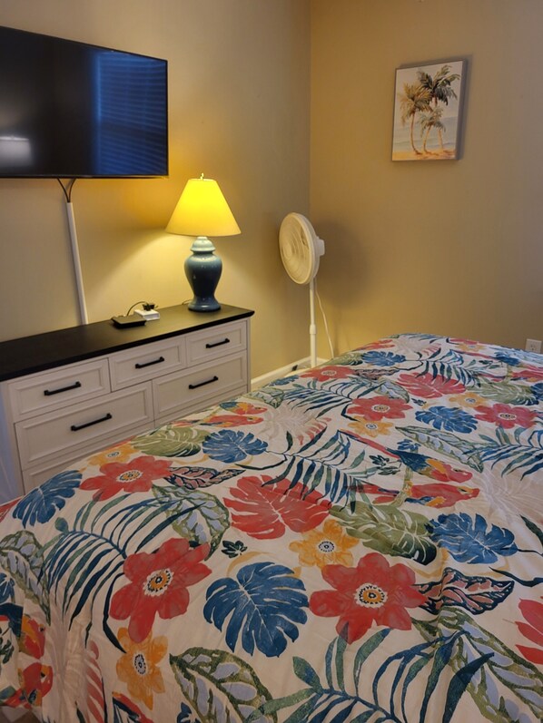 A bedroom with a floral bedspread, a bedside lamp, a fan, a TV on the wall, and a painting of palm trees.