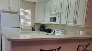 A kitchen with white cabinets, a refrigerator, a microwave, a coffee maker, and a kettle. There are three stools at the counter. - 2103 Palms, 2-bedroom condo, Great Fall Weekends are still available (Rehoboth Beach)
