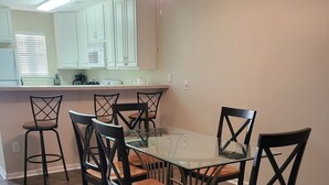A dining area with a glass-top table surrounded by six chairs with wooden frames and cushioned seats. The room has wooden flooring and white walls. - 2103 Palms, 2-bedroom condo, Great Fall Weekends are still available (Rehoboth Beach)