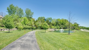 A paved pathway leading to a pond with a fountain, surrounded by lush green grass and trees. - Chic Family-Friendly Home in Riverside: 7 Mi to KC (Riverside)
