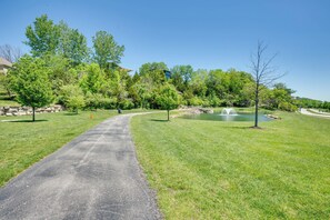 A paved pathway leading to a pond with a fountain, surrounded by lush green grass and trees. - Chic Family-Friendly Home in Riverside: 7 Mi to KC (Riverside)