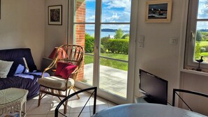 A room with a sofa, a wicker chair, a television, and a sliding glass door leading to a balcony with a view of the sea and greenery. - Nest with beautiful garden and sea view (Dinard)