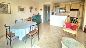 A dining area with a round table and chairs, a kitchen with wooden cabinets, and a wicker chair with red cushions. - Nest with beautiful garden and sea view (Dinard)