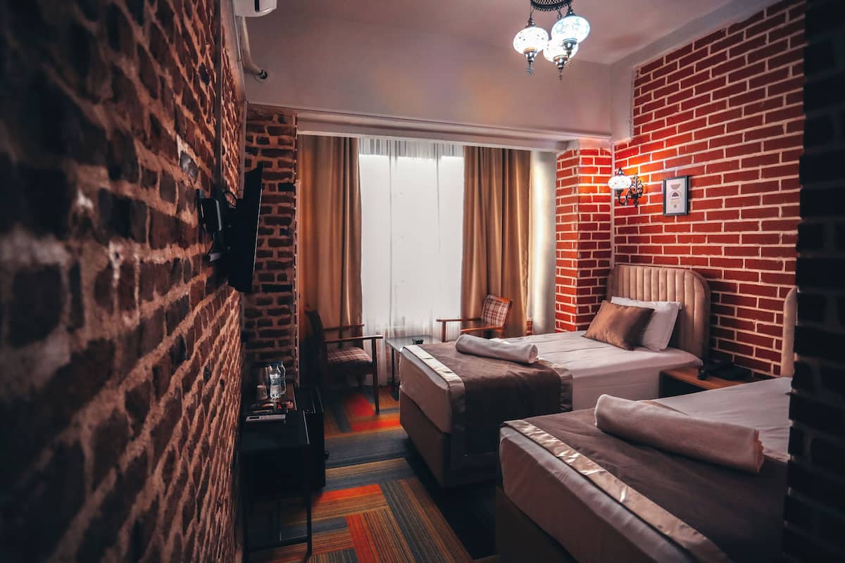 A hotel room with brick walls, two beds, a small table, a chair, and a window with curtains.