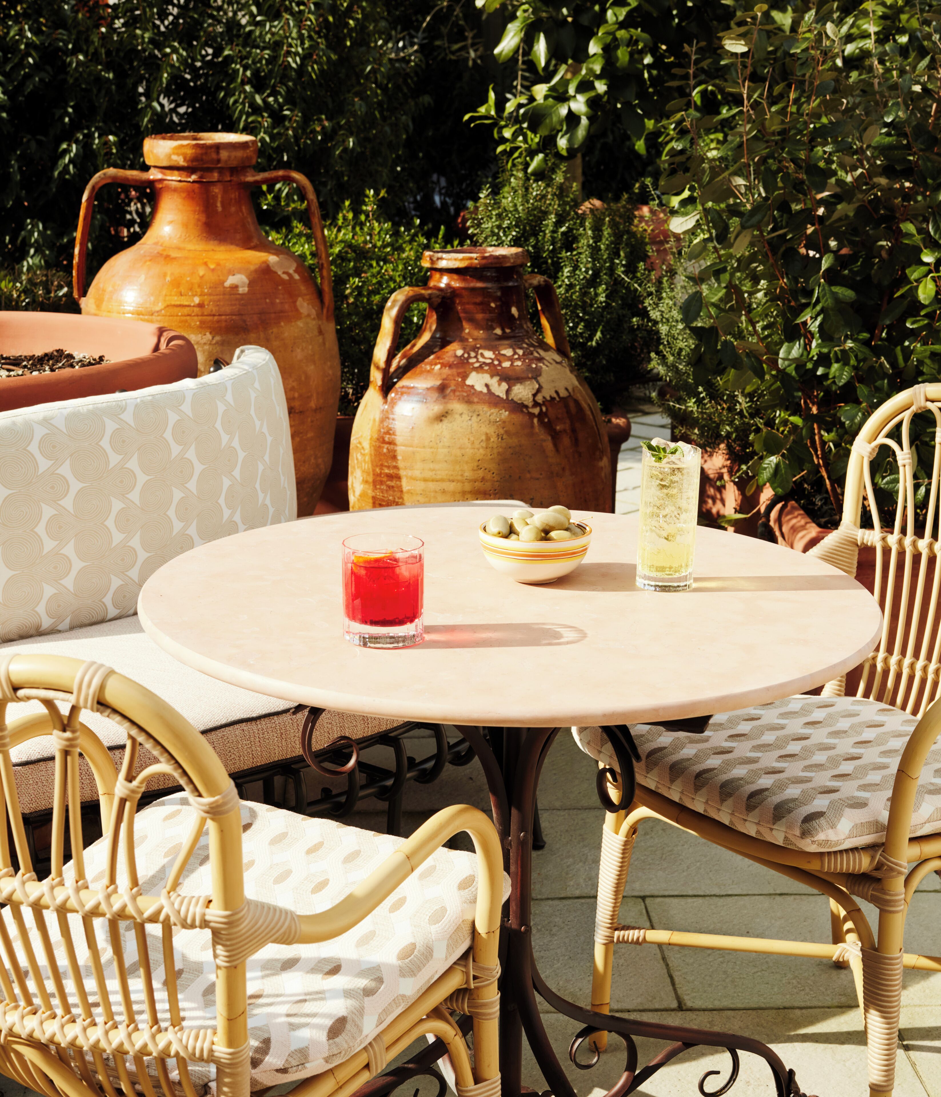 A round table with wicker chairs, a glass of red drink, a bowl of olives, and two large ceramic pots in a garden setting.