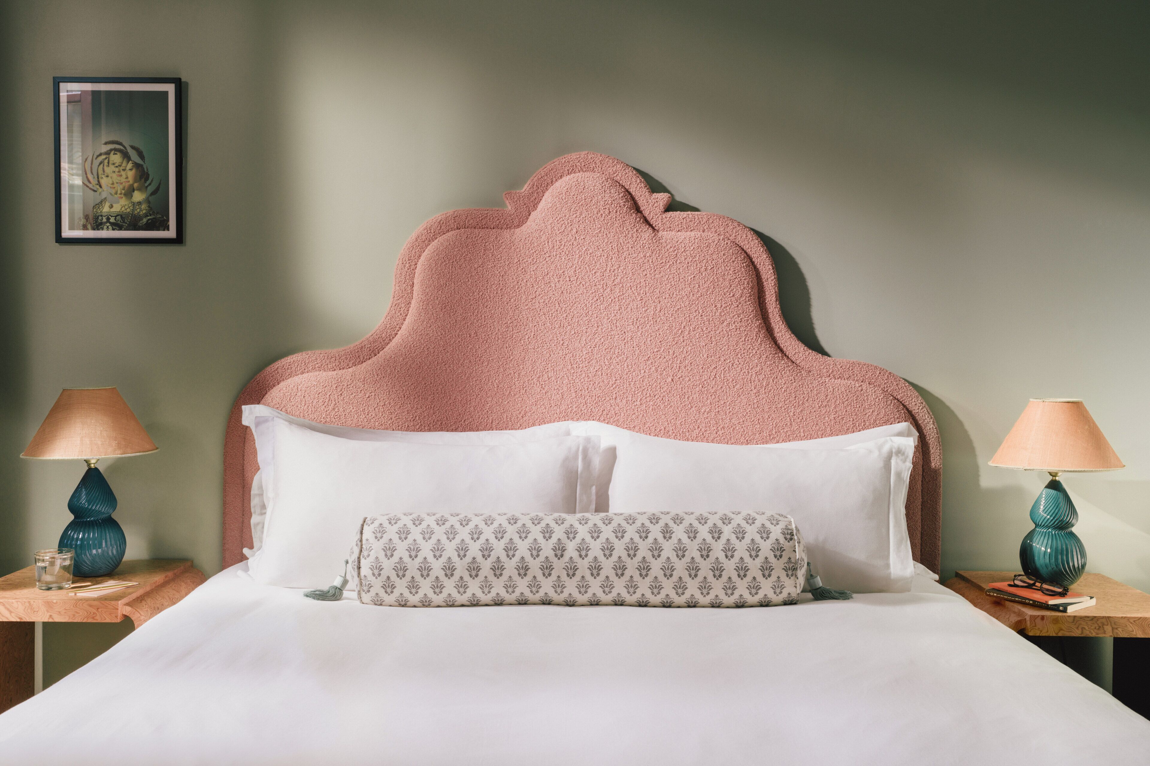 Un lit bien fait, avec une tête de lit rose, des oreillers blancs et un coussin à motifs. Il y a deux tables de chevet avec des lampes et un tableau encadré au mur.
