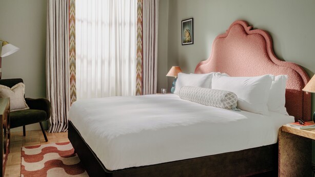 En pænt redt seng med hvide sengetøj, en rosa polstret sengegavl og et natbord med en lampe.