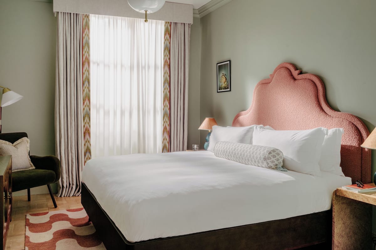 A neatly made bed with white linens, a pink upholstered headboard, and a bedside table with a lamp.
