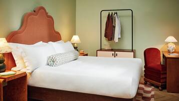 A neatly made bed with a wooden headboard, two bedside tables with lamps, a red chair, a wooden desk, and a clothing rack against the wall.