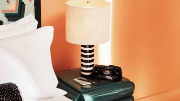 A neatly made bed with white linens, a black and white striped lamp, a black telephone, and a colorful abstract painting on the wall.