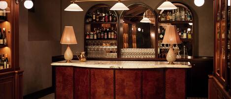 Zona de bar con una barra curva de madera, dos lámparas y un espejo que muestra botellas y vasos.