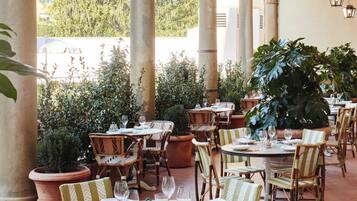 An outdoor dining area with round tables and wicker chairs, surrounded by large potted plants and columns.