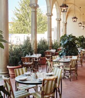 Zona pranzo all'aperto con tavoli rotondi e sedie in vimini, circondata da grandi piante in vaso e colonne.