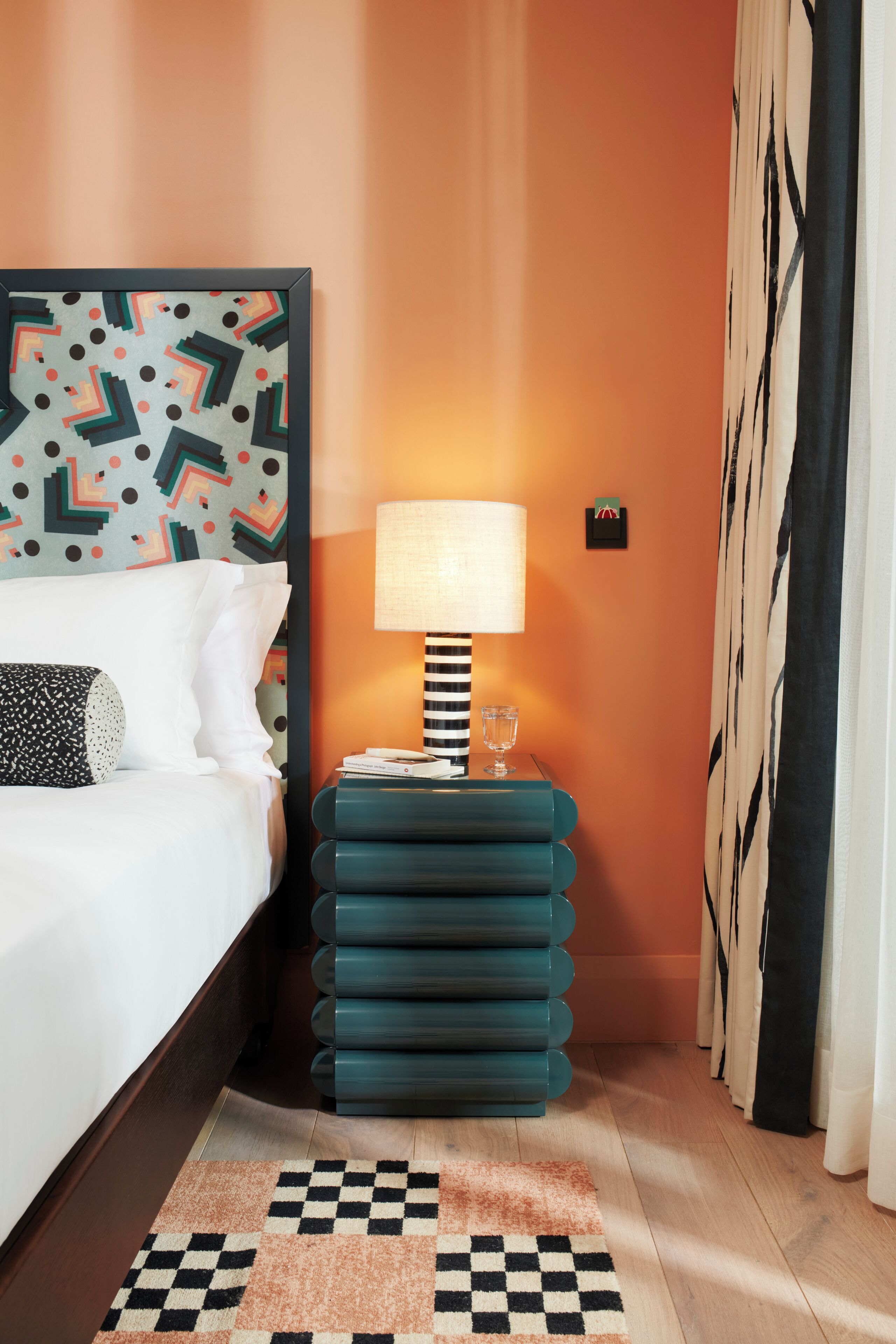 Ett hotellrum med en säng, en mönstrad sänggavel, ett turkosfärgat sidobord med en lampa, en rutig matta och en gardin.